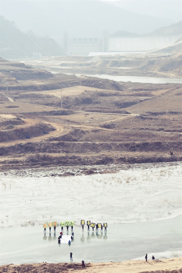 2016년 1월 환경운동연합 활동가들이 영주댐 해체를 주장하는 퍼포먼스를 벌이고 있다. 그 뒤로  금강마을이 보이고 그 너머에 영주댐이 서 있다. 