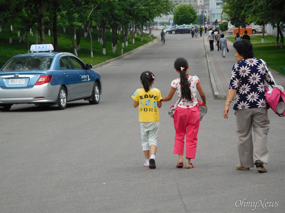  밝은 색 옷을 입은 북한 어린이들. 멀리 북한 택시가 보인다.