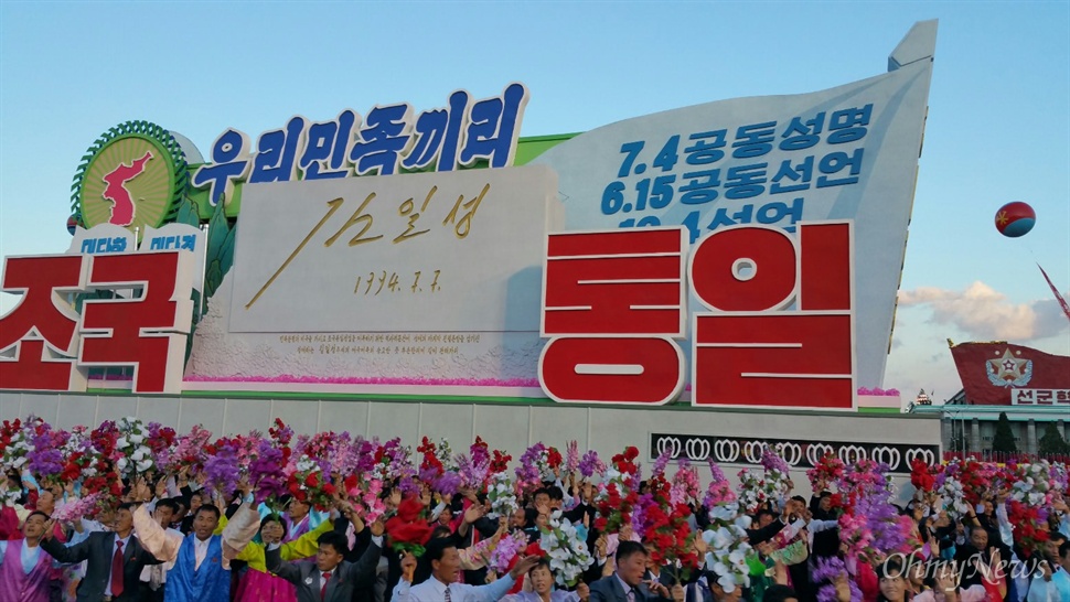 조선노동당 창건 70년 기념 열병식 현장. '우리민족끼리 조국통일'이라는 문구가 적혀 있는 구조물 아래서 열병식에 참석한 관중들이 꽃을 든 채 두 팔을 들어올리고 있다.