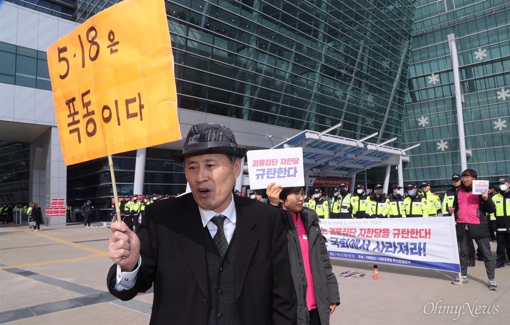 "5.18은 폭동이다" 주장하는 이 사람 21일 부산 벡스코에서 열린 자유한국당 부산·울산·경남·제주 합동연설회 행사장 입구에 "5.18은 폭동이다"라고 주장하는 한 시민이 지나고 있다. 뒤로 '5.18 망언' 김진태, 김순례, 이종명 의원 퇴출을 요구하는 부산 시민들의 현수막과 대비된다. 