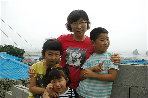 지오 어린이가 집 앞에서 엄마와 누나, 동생과 함께 기념사진을 찍으며 장난을 치고 있다. 담장 너머로 가거도 먼 바다가 보인다.