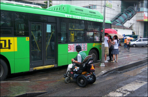 한 지체장애인이 저상버스차량에 오르지 못한 채 밖에서 저상버스차량에 오르는 비장애인을 바라보고 있다. 