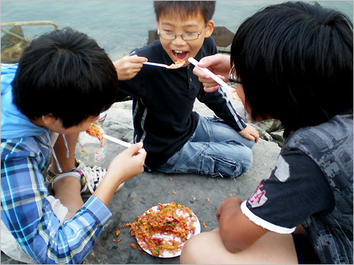 어린이 3명이 바닷가 제방에서 재첩비빔밥을 맛있게 먹으면서 맵다고 야단법석이다.