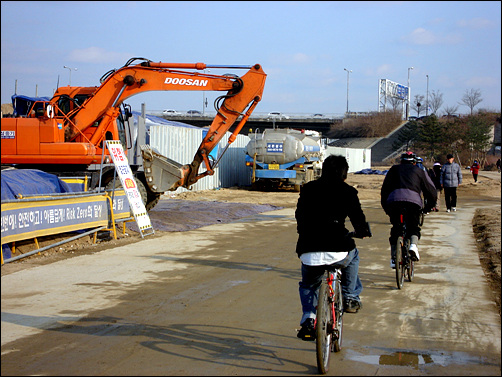 공사현장과 맞닿은 임시 자전거도로는 자전거족과 시민들로 붐빈다. 공사현장에서 포클레인이 튀어나온다.