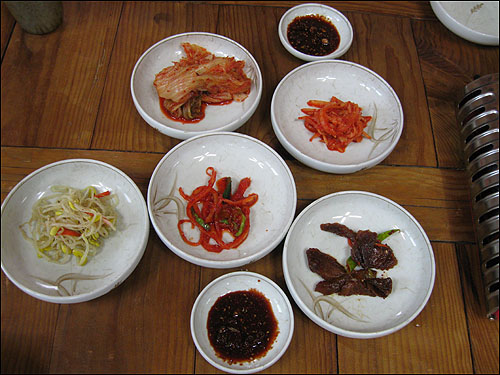 밑반찬으로 김치, 무채나물, 도라지무침, 콩나물, 무와 오이로 담근 오색장아찌와 함께 양념간장을 식탁 위에 올린다

