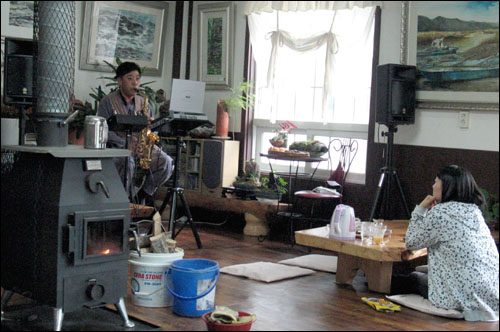 강금채 씨가 숲속 자신의 집 거실에서 색소폰을 연주해 보이고 있다. 그림작품이 즐비하고 장작난로가 놓인 거실에서도 예술적 감각이 느껴진다.
