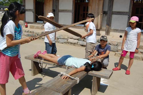 어린이들이 옥사 마당에 늘어놓은 형틀에 엎드려 곤장맞는 모습을 시연해 보이고 있다. 
