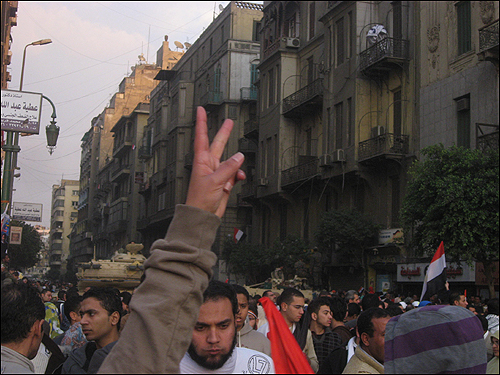 11일(현지 시각) 오후 이집트 수도 카이로의 타흐리르 광장에 시민들이 '무바라크 퇴진'을 요구하고 있는 가운데, 한 여성이 카메라 앞에서 승리의 브이(v)자를 그려 보이고 있다. 그 너머로 광장을 둘러싼 탱크가 보인다.
