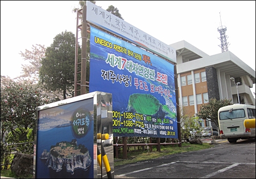  제주도청에 설치된 세계7대자연경관 대형 현수막. 그 앞쪽에는 해군기지가 건설되는 강정마을 인근 범선 사진이 있다. 