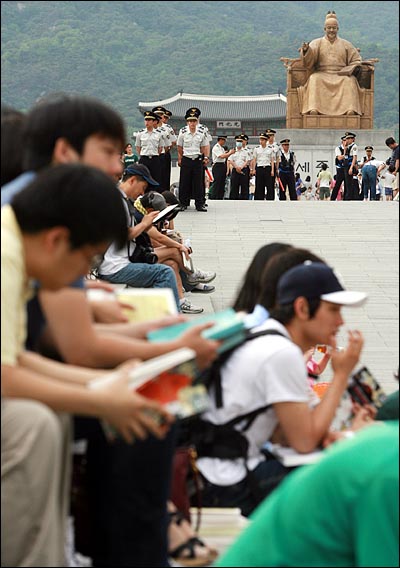  대학생들이 책을 읽는 가운데, 경찰들이 광화문 광장 입구에서 경비를 서고 있다.