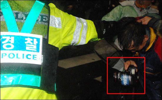 경찰에 머리채를 잡힌 사진기자의 손에 들린 후레쉬에는 파란 바탕에 흰 글씨로 'PRESS'가 찍힌 큼지막한 스티커가 붙어 있다.