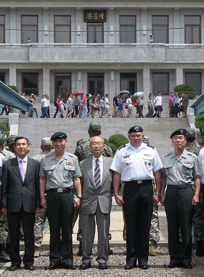 7월 27일 판문점에서 열린 정전협정 59주년 기념식에서 참석자들이 군사분계선 앞에서 기념촬영을 하고 있는 가운데 뒤쪽으로 판문각을 방문한 관광객들이 지나가고 있다. 왼쪽부터 국방부 임관빈 정책실장, 권오성 한미연합사 부사령관, 백선엽 장군, 제임스 D. 서먼 한미연합사령관, 이양구 유엔사 군정위 수석대표.