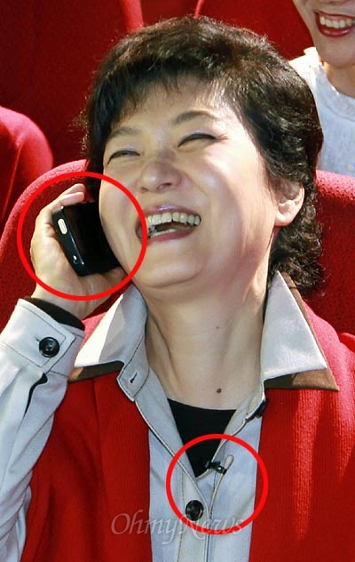 아이폰을 거꾸로 들고 전화통화 퍼포먼스를 하는 박근혜 후보. 옷에는 핀 마이크가 부착되어 있다.