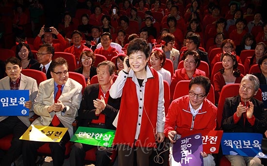 박근혜 후보가 아이폰을 거꾸로 든 채 자리에서 일어나 30대 여성과 통화하는 퍼포먼스를 하고 있다. 옆자리에 앉은 정두언 전 의원이 핀마이크가 연결된 무선송신기를 들고 있다.