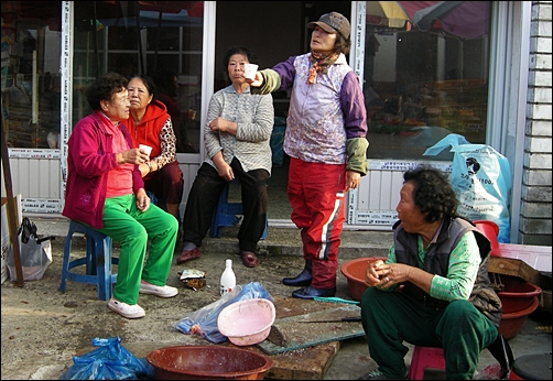 생선장수와 손님으로 보이는 할머니들이 막걸리를 마시고 있다. 
