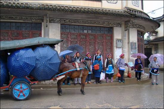 우리의 오일장처럼 매주 월요일마다 열리는 중국 윈난성 샤핑마을의 노천장터 앞에 백족(중국 소수민족)의 여인들이 집으로 돌아가는 마차를 기다리고 있다.(2011.7.18) 그 앞에 있는 마차에는 먼저 온 사람들이 저마다 우산을 쓰고 마주앉아 마차가 어서 떠나기를 기다리고 있다. 우리의 버스정류장처럼 기둥을 세워 마차를 탈 수 있는 표시를 해놓진 않았지만, 시장을 이용하는 사람들 사이에선 마차를 탈 수 있는 장소로 통하리라. 