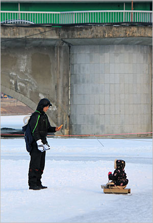 스마트폰으로 얼음썰매를 탄 딸아이를 찍고 있는 아버지. 