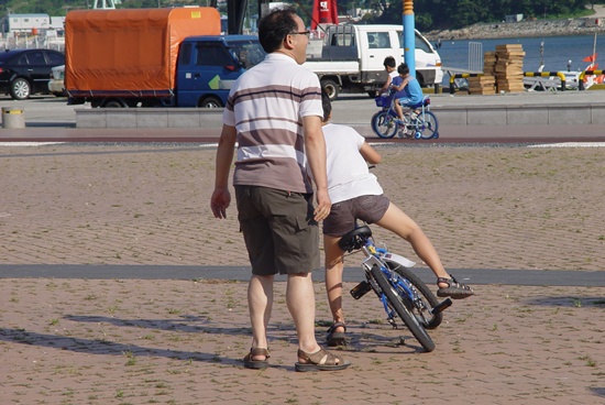 자전거 위에서 하염없이 넘어지는 아들과 무더위에 지친 아빠가 자전거 꽁무니에서 몸부림을 칩니다.