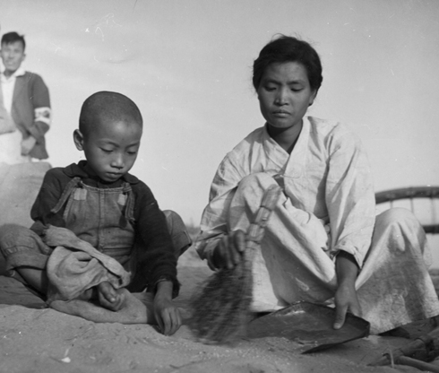 피난민 양곡배급소 바닥에 떨어진 낙곡을 모자가 빗자루로 쓸어 담고 있다(1950. 10.).
