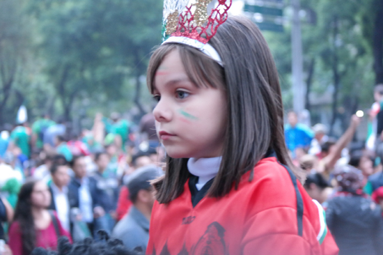 아빠의 등에 탄 소녀가 얼굴에 멕시코 국기 색칠을 하고 머리에 왕관을 쓰고 어리둥절한 축제를 즐긴다.