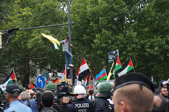 반이스라엘 시위대들이 베를린 쿠담 거리를 가득 메우고 있고 한 젊은 청년이 신호등에 올라가 깃발을 흔들며 구호를 외치고 있다.