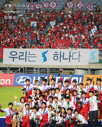  남과 북 여자축구 선수들이 함께 사진을 찍고 있는 뒤에 아리랑 응원단의 모습이 보인다. '우리는 하나다' 
