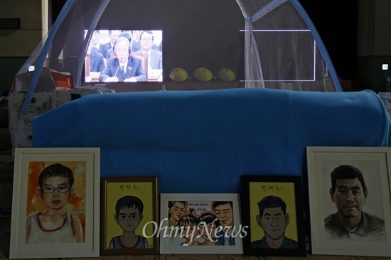 15일 실종자 권재근씨, 권혁규군 그림 너머의 진도체육관 대형 모니터에서 해양수산부 국정감사가 방영되고 있다.