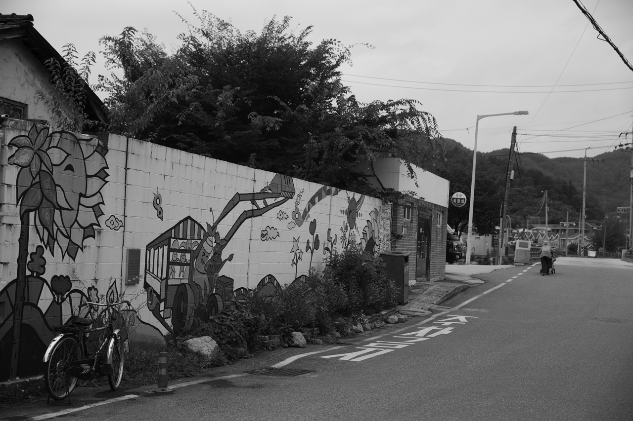 경기도 양평군 청운면 용두리에서 연화리 가는 길, 길가의 담벼락에 벽화가 그려져있고, 할머니 한 분이 걸어가고 있다.