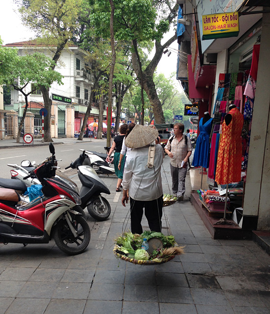  베트남 전통모자인 농을 쓰고 어깨에 채소들을 얹고 가는 상인의 뒷모습.