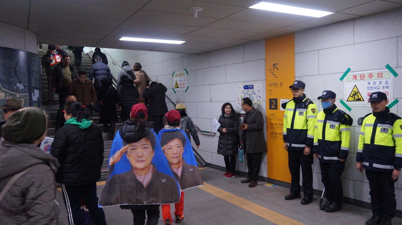 2번 출구 계단 앞 박근헤 대통령이 경례하는 사진이 포스팅된 망토를 둘러맨 사람들과 그앞을 지키는 경찰