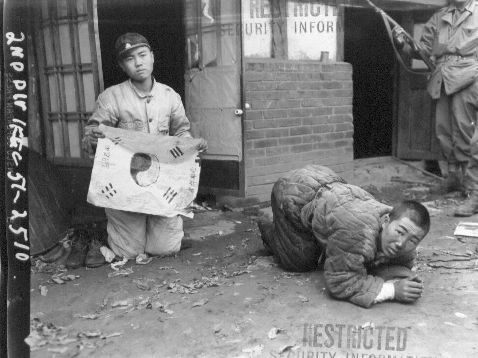  1951. 10. 21. 평양. 총구 앞에서 목숨은 구차하다. 학생은 태극기를 그려 들고, 인민군은 엎드려 살려달라고 애걸하고 있다.