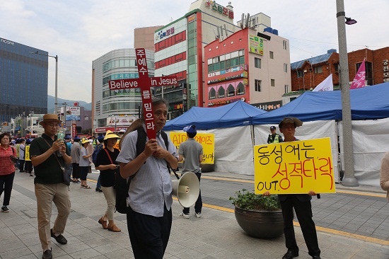 '동성애는 성적 타락'이라고 적힌 피켓을 든 사람 앞으로 십자가를 든 사람이 지나가고 있다.