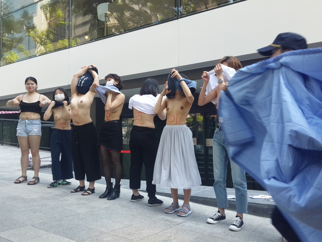 페이스북 사옥 앞에서 상의를 탈의하는 불꽃페미액션 회원들과 이를 급하게 제지하기 위해 이불로 가리러 달려오는 경찰들