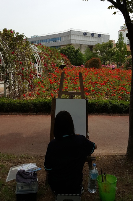 한 학생이 장미 동산을 배경으로 풍경화를 그리고 있다. 