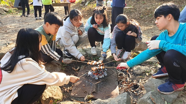 숲체험 프로그램에 참가한 초등학생들이 숯불을 중심으로 모여앉아 소시지와 떡을 굽고 있다.