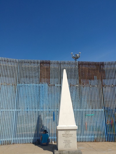 멕시코 영토의 최북단임을 알리는 기념비가 있는 곳에서 한 여성이 국경을 사이에 두고 그 너머 가족 혹은 지인과 대화하고 있다. 국경 철조망 위쪽에 미국측 감시 카메라가 보인다.
