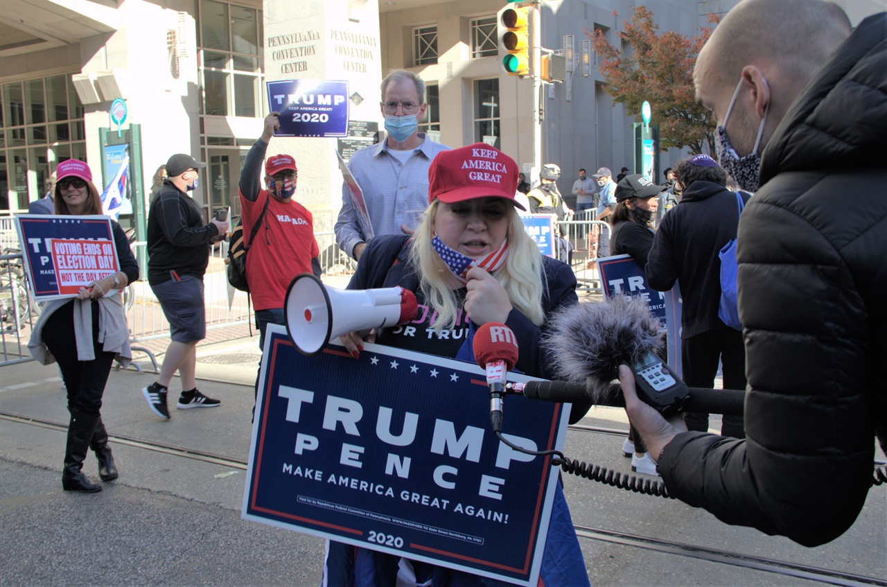 트럼프 대통령후보의 한 지지자가 개표소인 펜신베니아컨벤션센터앞에서 '미국을 다시 위대하게'라고 씌여진 모자를 쓰고 트럼프 대통령후보와 펜스부통령 후보의 손팻말을 들고 트럼프 후보 지지시위를 벌이며 한 언론과 인터뷰를 하고 있다.