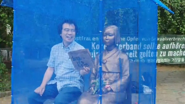 6월 29일 오후, 이우연 연구위원이 "요시코 15분 1.5엔"이 선명히 쓰인 피켓을 들고 베를린 평화의 소녀상 옆에 앉아 있다. 그는 다른 이와 대화하다가 웃음을 터뜨리기도 했다. (유튜브 화면갈무리)