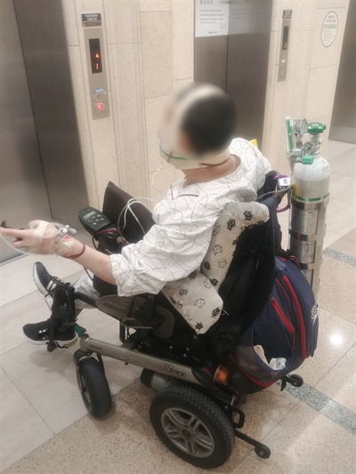 한 장애인이 전신마취 어금니 발치 후 진료를 보러 가고 있다. 병원에 알맞은 수동휠체어가 갖춰지지 않아 산소통을 메고 스스로 전동휠체어를 운전해 이동 중이다.