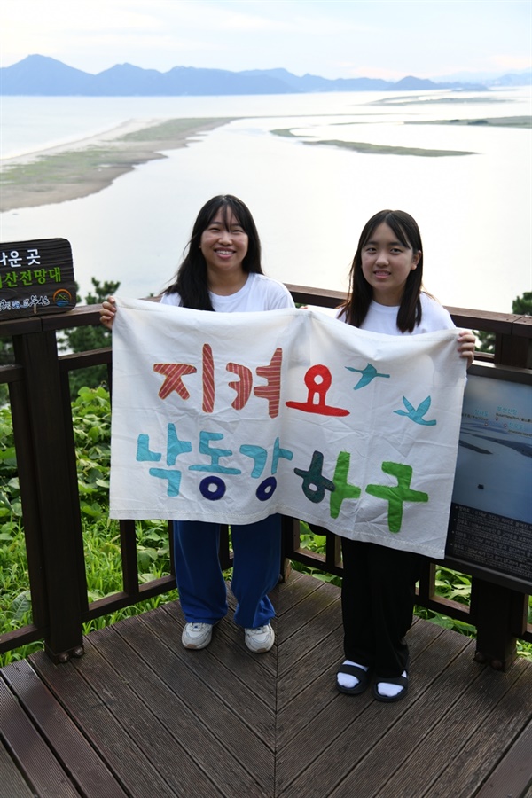 미래세대 낙동강 탐사대 두 소녀 대원이 든 현수막 뒤로 아름다운 낙동강 하구가 펼쳐져 있다. 그래도 한폭의 그림이다.  