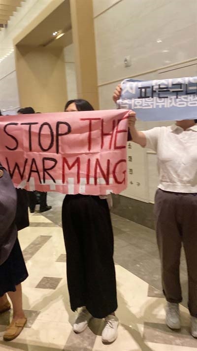 지난 9월 20일 고양 소노캄호텔에서 열린 DX KOREA 웰컴리셉션에서 전쟁없는세상 활동가들과 회원들이 기습시위를 펼쳤다. 한 참가자가 "Stop the WARming (전쟁을 멈춰라, 온난화를 멈춰라)"이라고 쓰인 현수막을 펼쳐 들고 있다.  