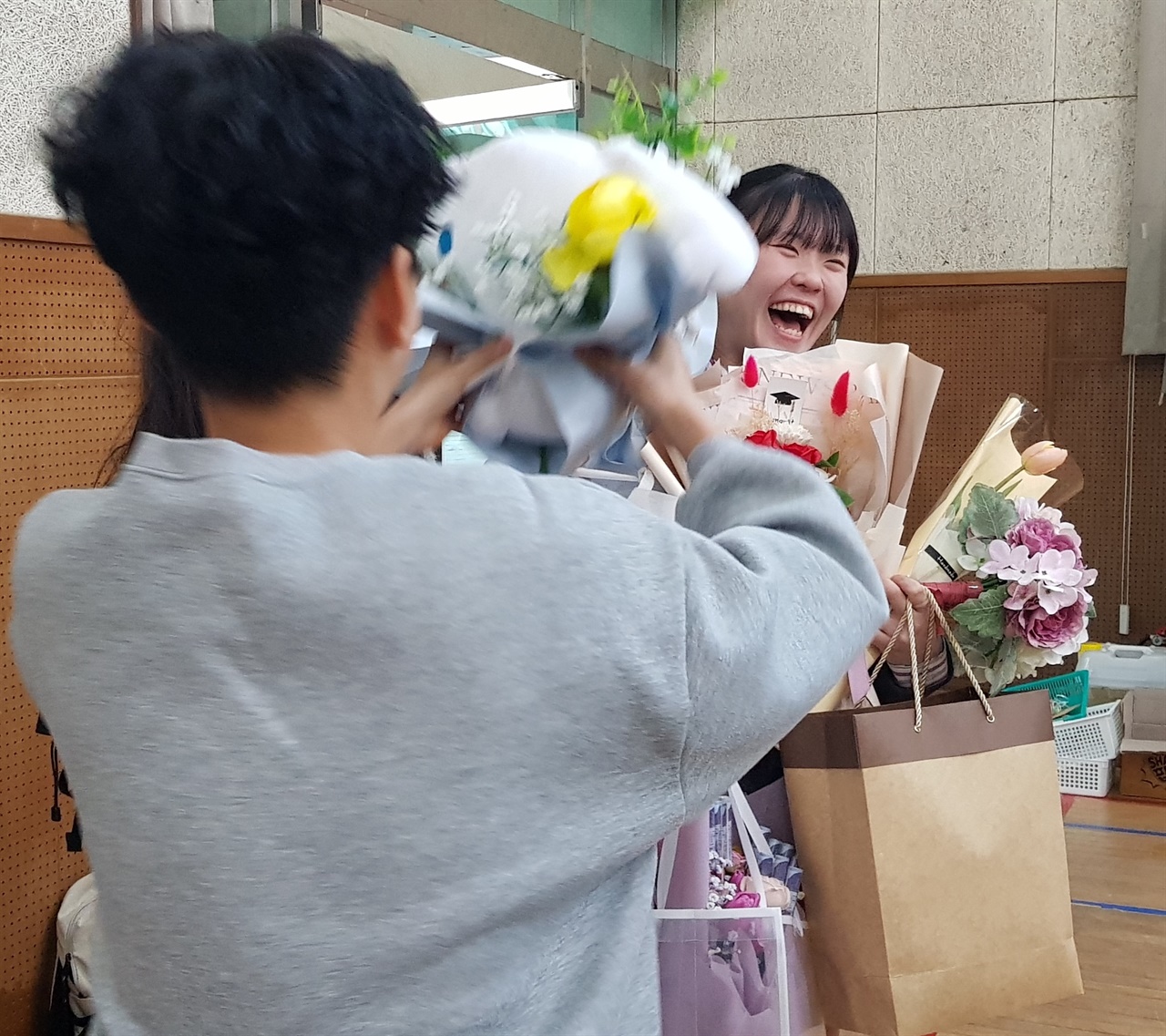 졸업식에서 여러 후배에게 꽃다발과 선물을 잔뜩 건네받은 학생이 활짝 웃고 있다.