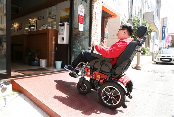 전동 휠체어를 탄 유청민 씨가 경사로를 설치한 ‘모두의 가게’로 들어가고 있다.