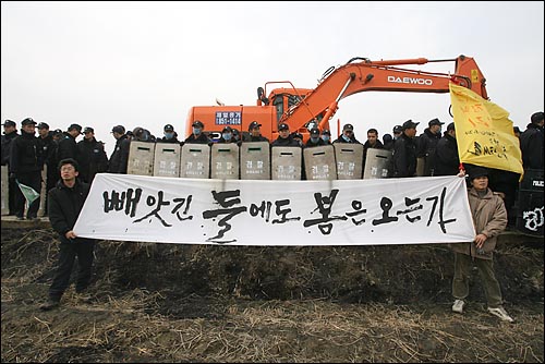 2006년 3월 15일 경기도 평택시 미군기지 확장예정지에서 농민들이 농사를 짓지 못하도록 국방부가 굴착기를 동원해 논에 구덩이를 파자 가수 정태춘(오른쪽)씨가 현수막을 들고 서 있다.